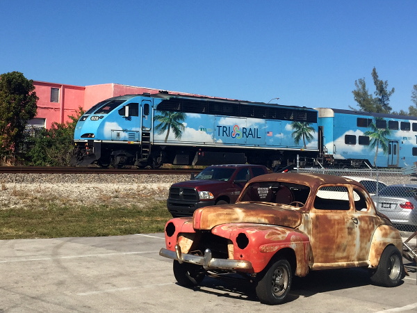 [Locomotiva  diesel-elettrica  Brookville BL36PH  nº 828  di
Tri-Rail  in transito  tra le  stazioni di  Fort Lauderdale  Airport e
Sheridan Street.  In  primo piano il relitto di una  Ford 1947.  Dania
Beach, 11 gennaio 2019.]